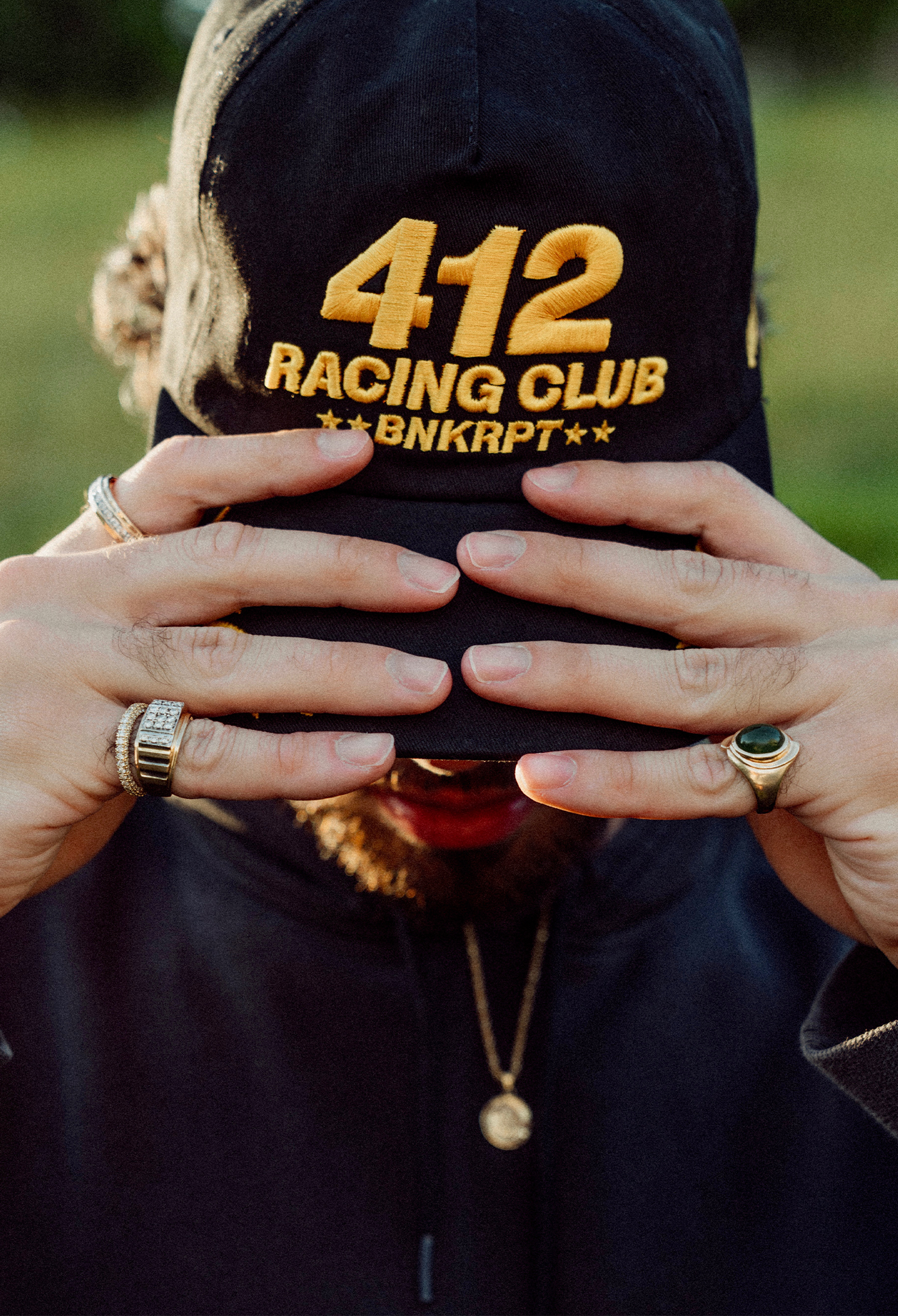 Person wearing a black cap with '412 Racing Club BNKRPT' text, hands covering face.